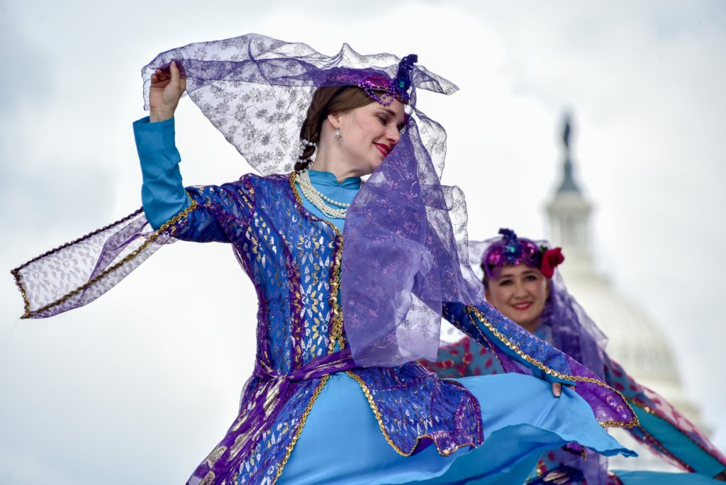 Person in elaborate teal and purple attire dancing with their scarf flowing around their face