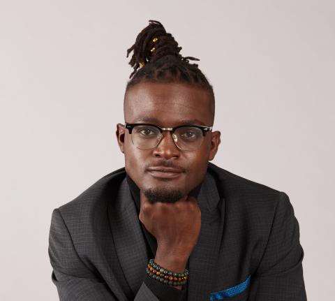 A black man wearing glasses with a suit on, with long dreadlocks pulled into a top bun, his head on his closed hand.